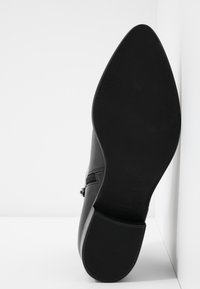 Semelle de bottine noire à bout pointu avec un talon texturé et une fermeture éclair latérale visible, positionnée à la verticale contre un fond d'angle blanc.