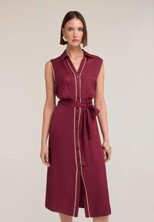 Frau mit kurzen braunen Haaren, die ein ärmelloses burgunderfarbenes Midikleid mit Knöpfen vorne und Taillenband trägt, dazu Ohrringe und Armbänder als Accessoires.