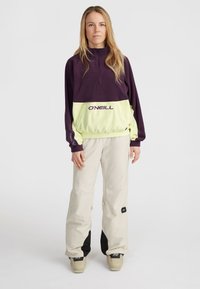 Paarse en limoen groene half-zip pullover met "O'Neill" logo; gecombineerd met lichtbeige broek met zwarte accenten en functionele details.