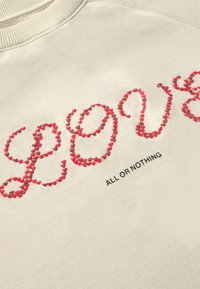 Beige Sweatshirt mit rotem gesticktem Text "LOVE" und einem dekorativen Muster, darunter schwarzer Text mit der Aufschrift "ALL OR NOTHING."