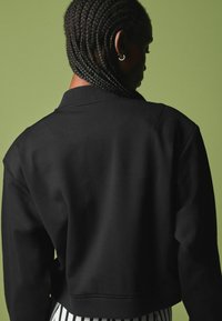 Sort cropped sweatshirt med ribbet krave og manchetter. Glat tekstur, vist fra ryggen mod en grøn baggrund.