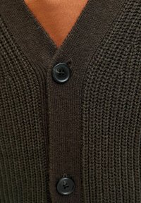 Tumši brūns adīts kardigāns ar V veida kaklu, kas rotāts ar teksturētu rakstu un divām melnām pogām priekšpusē. Zem tā redzams oranžs krekls.