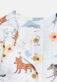 Koszulka z pikowanego bawełnianego materiału z białym tłem i kolorowymi wzorami zwierząt oraz kwiatów, wykończona brązowymi drewnianymi guzikami.