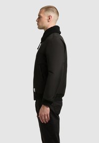 Schwarze Bomberjacke mit einem flauschigen Kragen, Reißverschluss und seitlichen Taschen. Glatte Textur, Rippbündchen und Saum.