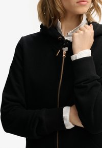 Felpa nera con zip e cappuccio regolabile, zip dorata e polsini a costine. Camicia bianca con colletto visibile sotto la felpa.