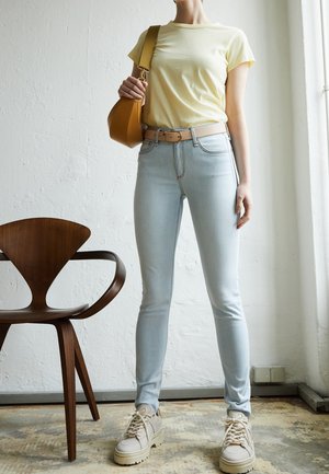 Person iført en gul T-shirt, lyseblå skinny jeans, beige bælte, hvide kraftige sneakers og holder en sennepsfarvet skuldertaske indendørs.