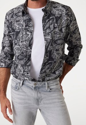 Muž nosící černo-bílé paisley vzorované košili s knoflíky přes bílé tričko a světle šedé džíny s rukama u boků.