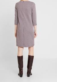 Femme portant une robe à motif pied-de-poule rose et noire arrivant au genou, avec des manches trois-quarts, des bottes haute jusqu'au genou de couleur brun foncé, debout de dos.