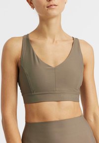 Mujer con top deportivo sin mangas de color verde oliva y mallas a juego de cintura alta, mostrada desde los hombros hasta el abdomen contra un fondo blanco.