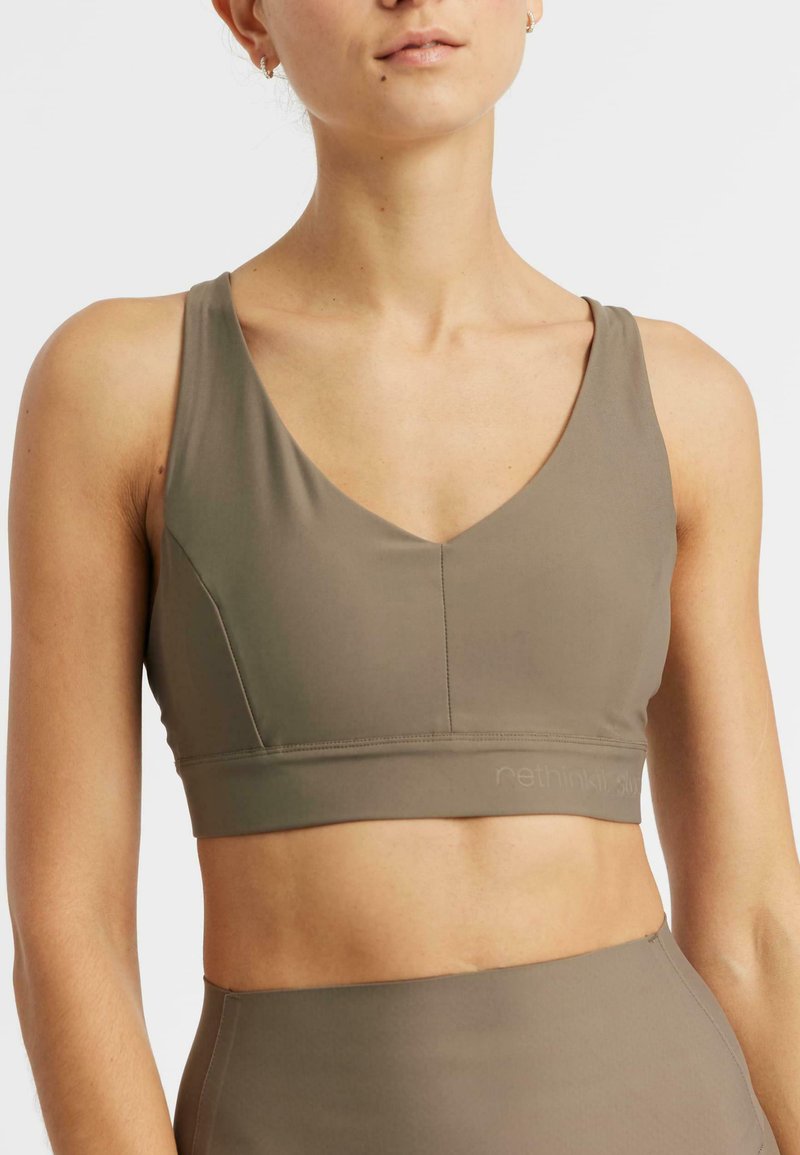 Mujer con top deportivo sin mangas de color verde oliva y mallas a juego de cintura alta, mostrada desde los hombros hasta el abdomen contra un fondo blanco.