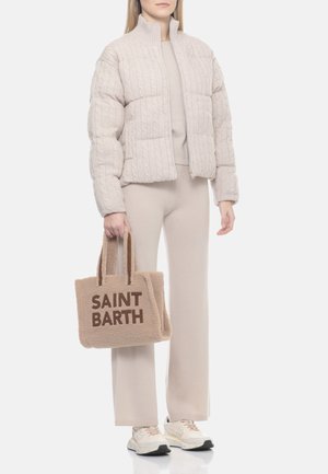 Donna che indossa una giacca a piumino beige a maglia a trecce, pantaloni larghi abbinati, sneakers e tiene una borsa tote testurizzata color cuoio con la scritta "SAINT BARTH".