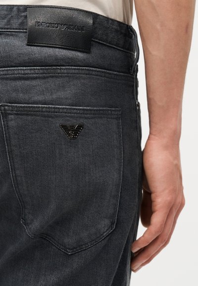 Τσέπη μαύρου τζιν με μαύρο λογότυπο αετού Emporio Armani και μαύρο δερμάτινο έμβλημα της μάρκας πάνω από αυτήν, συνδυασμένη με μπεζ πουκάμισο και εμφανές δεξί χέρι.