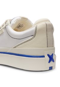 Κοντινή λήψη της φτέρνας λευκού αθλητικού παπουτσιού με υφή στη σόλα, μπλε ρίγα διακόσμηση, μαύρη ραφή και μπλε λογότυπο "X" στην ετικέτα της φτέρνας.