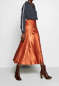 Blusa a pois navy con accenti bianchi, abbinata a una gonna in satin arancione fluida e stivali alti con tacco in vernice marrone effetto coccodrillo.