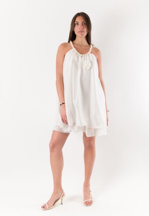 Giovane donna in piedi che indossa un vestito bianco senza maniche con cinghia di perle e dettaglio floreale, abbinato a tacchi beige su sfondo semplice.
