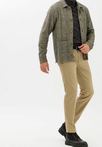 Męska koszula w kratę na guziki w kolorach zielonym i brązowym, zestawiona z beżowymi spodniami i czarnymi sznurowanymi butami. Charakteryzuje się teksturowaną tkaniną i luźnym krojem.