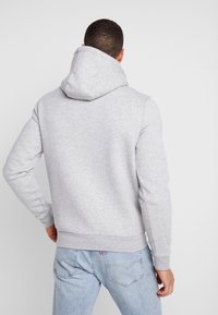 Sudadera gris con capucha amplia, puños y dobladillo acanalados, hecha de una tela de algodón texturizada. Sin logotipos ni patrones visibles.