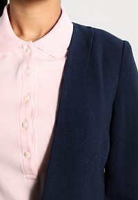 Veston bleu marine avec une texture subtile, associé à une chemise rose clair à col avec boutons. Gros plan sur les détails du tissu et du col.