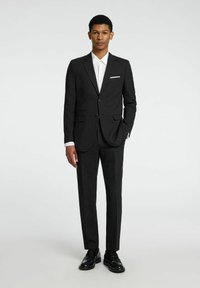 Traje gris oscuro de corte slim, con dos bolsillos frontales y un pañuelo blanco en el bolsillo, combinado con una camisa de vestir blanca y zapatos negros.