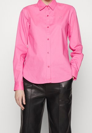 Kobieta ubrana w jaskraworóżową koszulę zapinaną na guziki z długimi rękawami oraz czarne skórzane spodnie, stojąca na tle jednolitego tła.