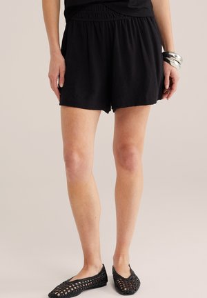 Žena v voľných čiernych šortkách, čiernych pletených balerínkach a masívnych strieborných náramkoch na neutrálnom pozadí.