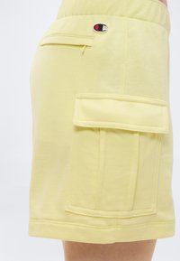 Κίτρινες βερμούδες με μεγάλο πλαϊνό τσεπάκι cargo και μικρή πίσω τσέπη με φερμουάρ, με λογότυπο Champion κοντά στη μέση.