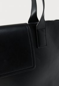 Μαύρη δερμάτινη τσάντα με λείο ύφασμα, με δομημένο σχεδιασμό, κοντά χερούλια και κλείσιμο με φερμουάρ. Μινιμαλιστικές λεπτομέρειες κατά μήκος των ακρών.