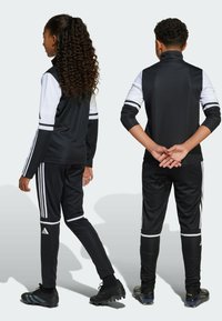 Czarny sportowy dres z wysokim kołnierzem, białymi panelami na ramionach i trzema białymi paskami na spodniach. Teksturowany, sportowy materiał.