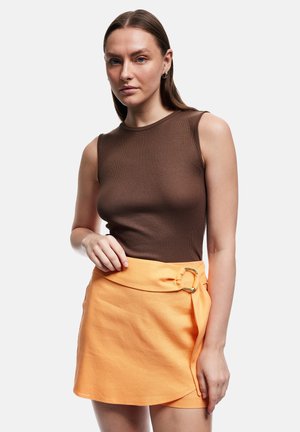 Kvinde med glat brunt hår iført en brun ærmeløs top og orange nederdel med høj talje og guldlås, stående mod en ensfarvet baggrund.