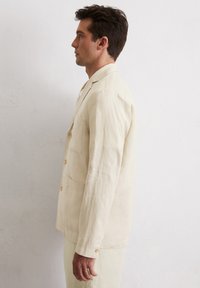 Hellbeige Leinenjacke mit einer einzelnen Brusttasche, drei Knöpfen und einem lässigen Schnitt, präsentiert an einem Modell im Profil.