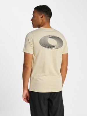 Mann trägt beige kurzärmeliges T-Shirt mit schwarzem, kreisförmigem Sonnenstrahl-Design auf dem Rücken, kombiniert mit schwarzen Hosen, blickt nach links vor weißem Hintergrund.