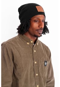 Black knitted beanie with a leather patch and a brown corduroy shirt featuring a small logo and golden buttons.