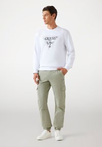 White sweatshirt with a triangular "GUESS" logo, paired with light green cargo pants featuring elastic cuffs and multiple pockets, worn with white sneakers.