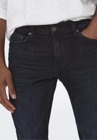 Jeans de mezclilla azul oscuro con un diseño ajustado, que cuentan con bolsillos frontales y acentos de botones de metal. La tela tiene una ligera textura y brillo.
