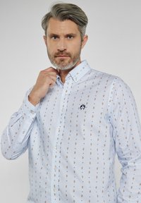Błękitna koszula z guzikami, z białym i złotym wzorem paisley, z małym haftowanym logo na piersi i usztywnionym kołnierzykiem.