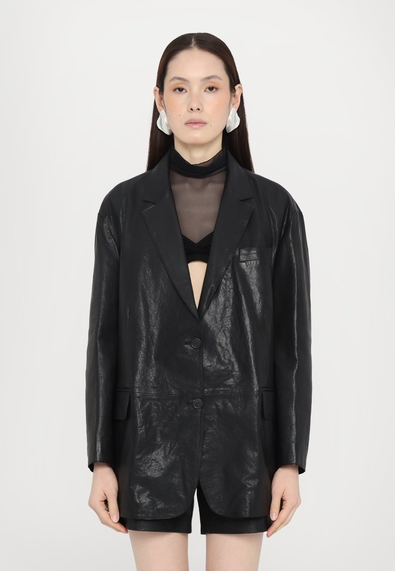 Blazer din piele neagră cu o textură creponată, închidere cu un singur nasture și două buzunare frontale. Purtați peste un top negru transparent.