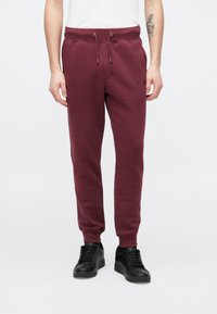 Μπεζ φόρμες μπορντό με ελαστική μέση και κορδόνι, με πλαϊνές τσέπες και μανσέτες ριμπ, κατασκευασμένες από απαλό, υφασμένο ύφασμα.