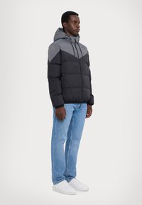 Giacca a vento nera e grigia con cappuccio, texture trapuntata, tasche con zip e design a pannelli a contrasto. Indossata con jeans azzurri chiari e sneakers bianche.