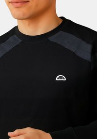 Maglietta nera a maniche lunghe con texture a coste, dotata di pannelli sulle spalle grigi tono su tono e un piccolo logo bianco sul petto.