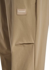 Beige techwear bukser lavet af glat stof, med et diskret Hummel-logo mærke på forsiden og en løstsiddende, afslappet pasform.