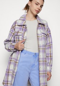 Manteau en laine à carreaux violets et blancs avec des accents marron, doté de grandes poches et de boutons-pression, porté par-dessus un haut gris clair ajusté.