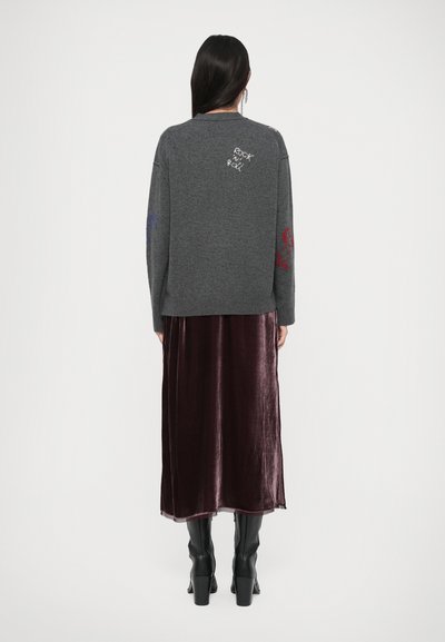 Grijze trui met blauwe en rode geborduurde ontwerpen op de mouwen, gecombineerd met een lange bordeauxrode fluwelen rok en zwarte kniehoge laarzen.