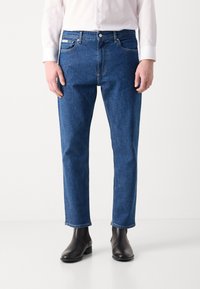 Jeans de mezclilla azul con corte recto, que presentan un diseño clásico de cinco bolsillos, costuras doradas y cierre de cremallera. Llevados con botas negras de tobillo.