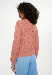 Roze gebreide trui met een open rug en sleutelgatdetails, voorzien van een kantpatroon en een geribde zoom. Gepaard met lichtblauwe denim broek.
