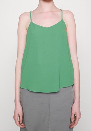 Femme portant un haut vert ample sans manches avec des bretelles fines et une jupe grise, se tenant devant un fond clair et uni.