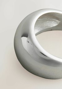 Sølvfarvet metalring med en glat, reflekterende overflade; rundet, bølgende form uden tydelige mønstre eller indgraveringer.