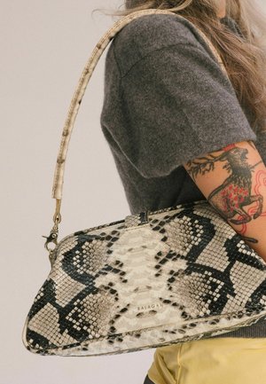 Persona con braccio tatuato che indossa una maglia grigia e porta una borsa beige e nera con motivo di pelle di serpente, con tracolla curva sulla spalla.