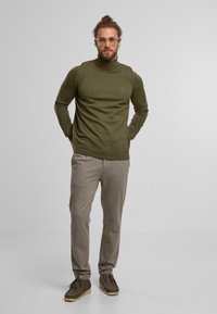 Olivgrüner Strickrollkragenpullover, schlanker Schnitt, gerippte Bündchen, kombiniert mit hellbeigen Hosen und braunen Schnürschuhen, stehend vor einem neutralen Hintergrund.