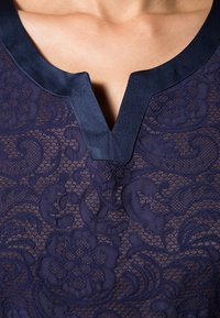 Námořnická modrá část oděvu s texturovaným květinovým vzorem a výstřihem do V. Obsahuje hladký saténový lem podél výstřihu.