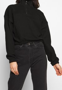 Black fleece pullover with high collar, half zip, elastic waistband, and cuffs. Paired with dark denim jeans, overall relaxed fit.